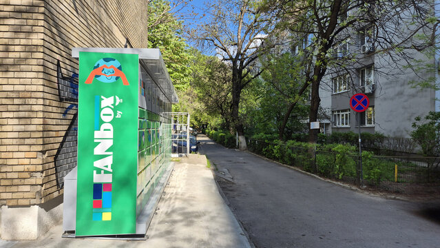 A bright green FANbox automated parcel locker stands on a sidewalk next to a brick building and a narrow tree-lined street. Picture taken on April 23, 2026 in Bucharest, Romania.
