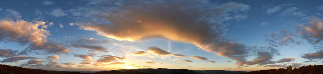 Fototapeta premium Stunning sunset fills sky with rich hues of blue and orange. Dramatic clouds with hints of pink and gold spread across sky. Silhouettes of hills in foreground add depth to breathtaking evening scene.