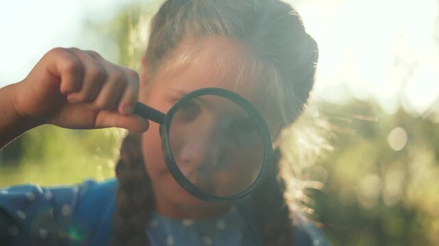 Curious child looks through magnifying glass outdoors. Girl explores nature in sunny park. Kid with braid holds magnifying lens near eye. Child discovery and exploration in warm sunlight.
