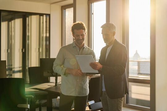 Two men in an office having a business meeting at sunset, using a tablet