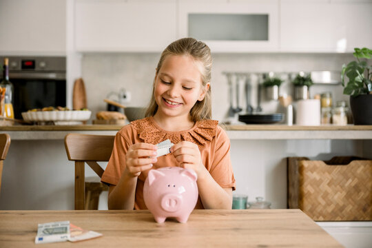 Girl saving money in piggy bank at kitchen table and smiling
