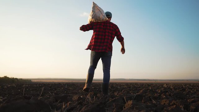 Farmer carrying sack walks across plowed field at sunset. Man in boots strides over soil. Farmer hauls heavy bag on shoulder. Rural agriculture work on farmland. Plowed earth stretches to horizon.
