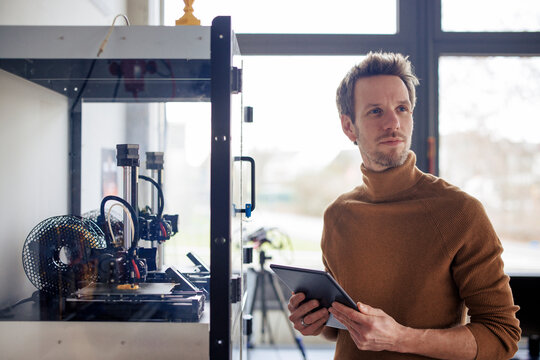Thoughtful engineer holding tablet PC next to 3d printing machine at workshop