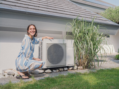 Smiling woman kneeling by thermal heat pump in house garden in summer