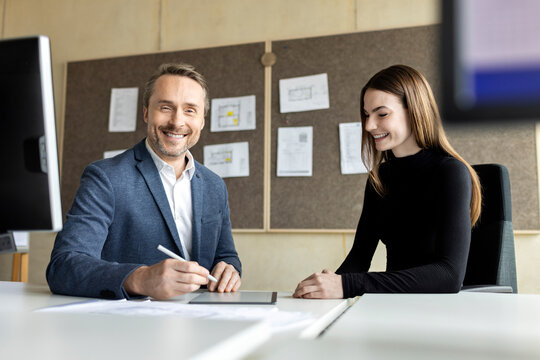 Smiling businessman with tablet PC on desk by trainee in office