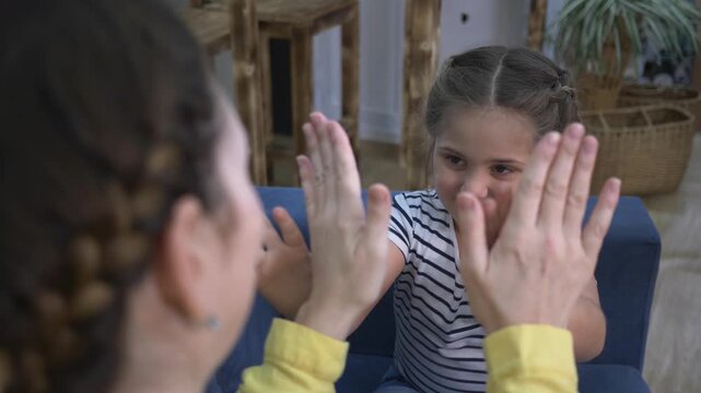 Mother and child playing clapping hand game on sofa. Girl smiling while playing with mom at home. Happy kid claps hands with parent. Family bonding through play indoors. Child enjoys clapping game.