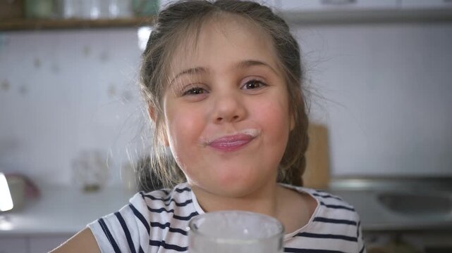 Little girl drinks milk from clear glass in kitchen. Cute child lowers glass from face. Funny milk mustache stays on upper lip. Happy girl shows big smile. Young child loves healthy milk drink.
