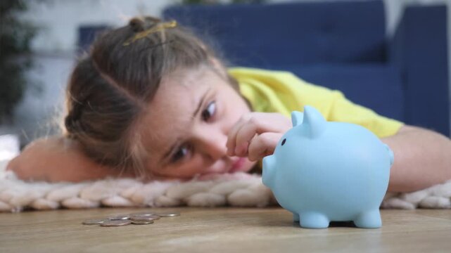 Little girl lies on wooden floor saving money. Cute child looks at blue piggybank. Kid holds small metal coin in hand. Hand puts coin into money box. Girl learns finance. Child plans investment.