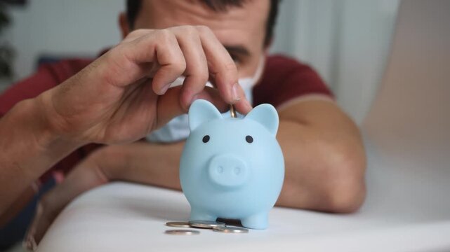 Man in face mask saving coins in piggy bank. Saving money during pandemic crisis. Man counts coins near blue piggy bank. Financial saving and budget planning at home. Coin deposit into piggy bank.