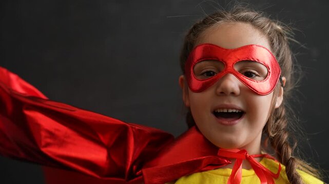 Happy child in superhero costume smiles with joy. Girl wears red mask and cape flowing in wind. Superhero kid laughs and plays hero. Child in red cape enjoys pretend play. Happy girl with mask grins.