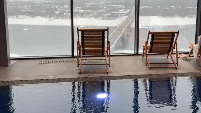 Wooden sun loungers line the spa center pool deck before expansive panoramic windows revealing stunning city skyline views and distant horizon