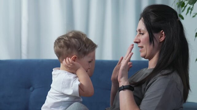 Mother playing peekaboo with toddler son. Mom and child clap hands on sofa. Mother plays with baby boy at home. Parent and toddler bonding together. Mom teaches kid hand game on couch indoors.
