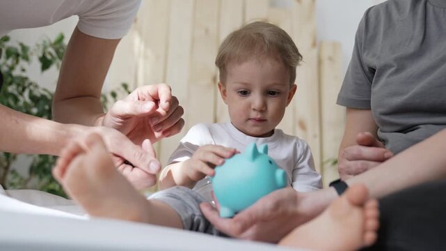 Toddler sits on bed with family. Child puts coin into light blue piggy bank. Parent holds bank for toddler. Family teaches early finance lessons. Saving money starts young. Coin drops in bank.