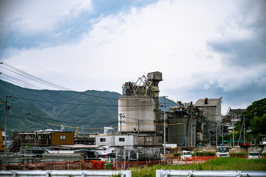 山並みを背景に稼働する巨大なセメント工場のプラントと手前に広がる建設現場の風景。 / A large cement plant operating against a mountain backdrop with a construction site in the foreground.