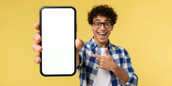 Excited man points to blank smartphone screen. Guy shows mobile phone display with copy space for app ad. Positive teen promotes new tech on yellow background.