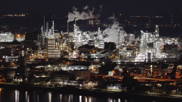 Aerial view of the BASF chemical plant glowing brightly at night, with white steam rising against the dark sky and reflections in the river below, Ludwigshafen am Rhein, Rheinland-Pfalz, Germany.