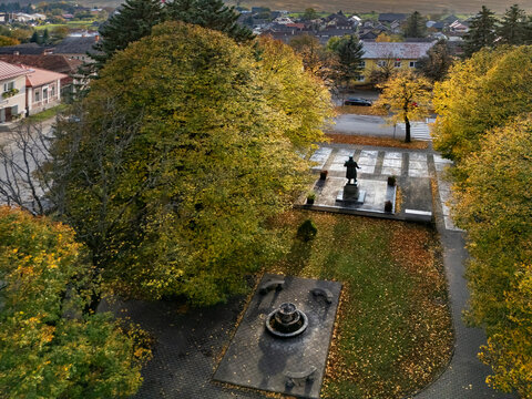 Aerial view of a serene autumnal park, where golden and green foliage frames a solemn statue and a dark modern fountain, Mosovce, Zilina Region, Slovakia.