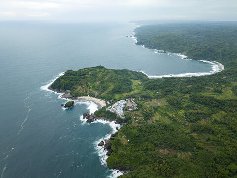 Aerial view of Jungwok Beach where the turquoise sea meets the verdant cliffs under a cloudy sky, Gunungkidul Regency, Special Region of Yogyakarta, Indonesia.