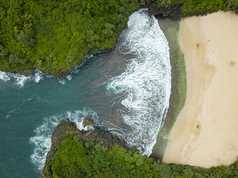 Aerial view of the Sedahan Beach where turquoise waters meet the shore with foamy waves, framed by lush green cliffs, Gunungkidul Regency, Indonesia.