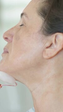 Vertical video: Rolling woman in light top moving quartz roller along jaw and neck at home for skin
