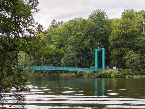 Turquoise Kłosowski suspension bridge over Raduń Lake in Wałcz, West Pomerania, Poland, surrounded by trees and water.