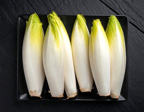 Close-up of six pale, oblong leafy vegetables arranged neatly on a dark square plate against a textured black surface