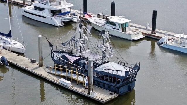 Aerial view of a dark, tattered pirate ship replica docked alongside modern yachts in a serene marina, Fernandina Beach, Florida, United States.
