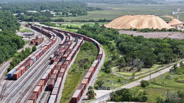 Aerial view of rows of freight trains snake through green trees, contrasting with the immense golden piles of phosphate, Fernandina Beach, Florida, United States.