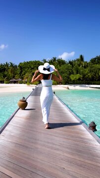 Rear view of a elegant woman in white dress and hat walks down a pier towards a tropical island in the Maldives