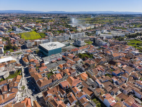Aerial view of a vibrant urban tapestry woven with ancient terracotta roofs and a striking modern teal structure, bathed in the bright Portuguese sun, Ilhavo, Aveiro, Portugal.