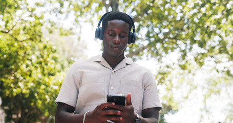 Fototapeta premium Black man, phone and music at park with headphones, streaming or listen to sound with low angle in summer. African person, scroll and outdoor with mobile app, texting or audio subscription in nature