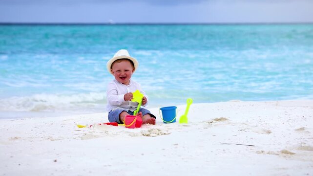 A cute baby or toddler boy plays with colourful sand toys on a tropical beach with turquoise sea