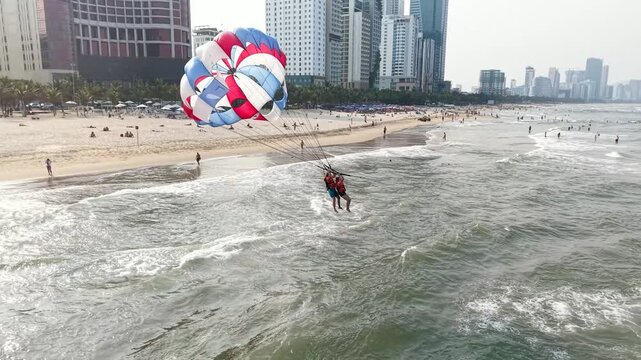 People enjoying parasailing activity at my khe beach, da nang, vietnam, preparing to launch into the sky over the ocean, surrounded by a lively beach scene with a modern city skyline backdrop