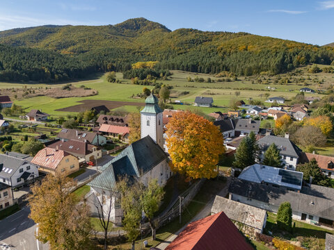 Aerial view of a village church with a green spire and a brilliant autumn tree, amidst homes and dark green hills, Klastor pod Znievom, Zilina Region, Slovakia.