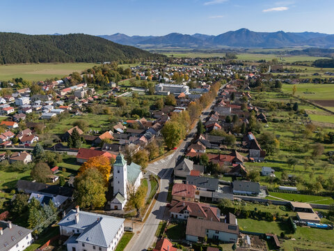 Aerial view of a serene village with a distinctive white church spire, vibrant autumn foliage, and a winding road, framed by distant mountains, Klastor pod Znievom, Zilina Region, Slovakia.