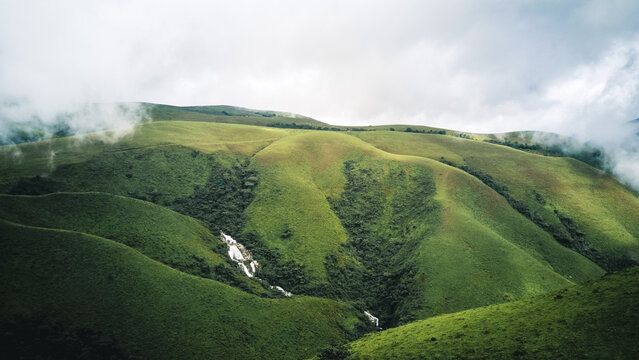 Aerial view of rolling emerald hills kissed by soft, ethereal mist, with a shimmering waterfall carving a path through the vibrant landscape, Obudu, Cross River, Nigeria.