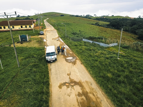 Aerial view of a white van and people on a muddy road, with rolling green hills and a brown-roofed building under an overcast sky, Obudu, Cross River, Nigeria.