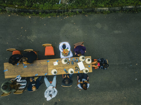 Aerial view of a long wooden table set for a communal meal, surrounded by people gathered on chairs, contrasting sharply with the dark asphalt road, Obudu, Cross River, Nigeria.