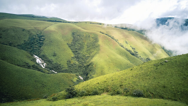 Aerial view of lush, undulating green hills kissed by soft white clouds, revealing a cascading waterfall amidst verdant slopes, Obudu, Cross River, Nigeria.
