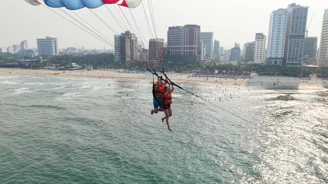 Two people attached to a parasail experiencing an exhilarating water sport adventure over the turquoise sea, offering a unique aerial view of the bustling beach and modern city