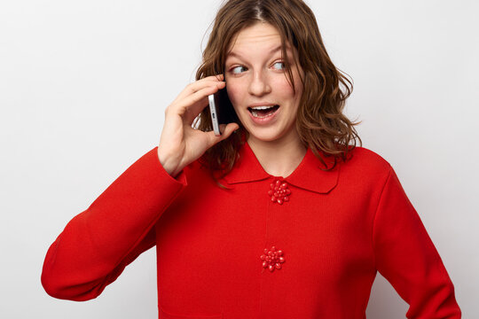 Young woman in red sweater talks on smartphone with surprised expression. Isolated on white background. Concept of communication, technology, emotions, and casual lifestyle.