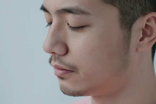 Man with closed eyes in profile, natural skin texture visible. Gentle facial features, light stubble on chin and upper lip. Clean, calm, and serene expression. Minimal background focuses on portrait.