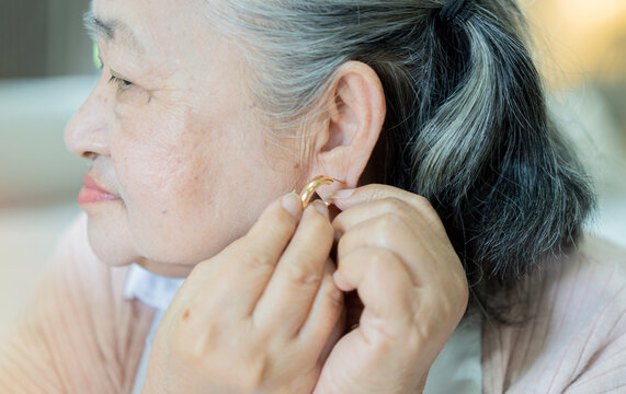 Close-up side view of Asian senior woman putting on earrings in a comfortable living space.