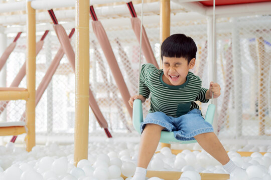 Little Asian boy having fun on a swing surrounded by white ball pits in the playground.