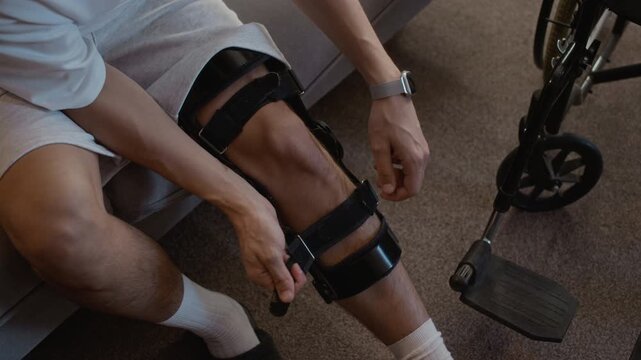 High angle shot of anonymous young man sitting on sofa and applying black orthopedic brace on injured leg