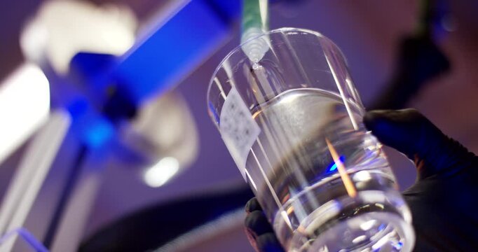 Low angle view of a robotic arm dispensing a green liquid sample into a glass of clear water in a lab