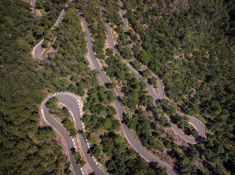 Top-down aerial view of a serpentine mountain road with multiple hairpin switchbacks winding through a dense green forest on a sunny day