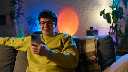 Fototapeta na wymiar Smiling man relaxing on sofa while using smartphone in cozy interior with colorful lighting. Concept of online shopping, digital lifestyle, ecommerce for retail and home services.
