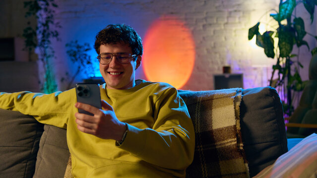 Smiling man relaxing on sofa while using smartphone in cozy interior with colorful lighting. Concept of online shopping, digital lifestyle, ecommerce for retail and home services.