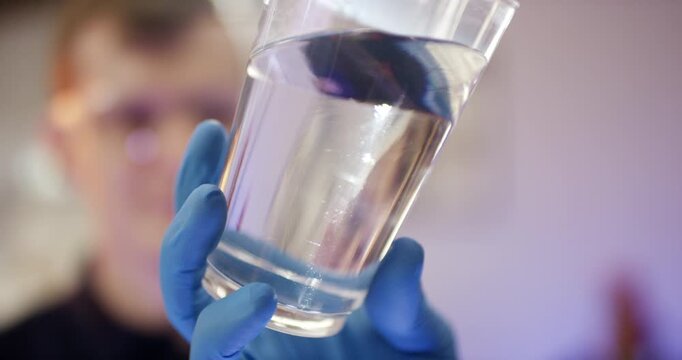Close up of a person wearing blue nitrile gloves holding a transparent plastic cup filled with clear liquid